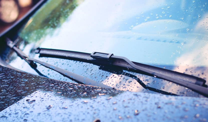 Close-up of windscreen wipers on a wet car windshield with raindrops, reflecting a blue sky.